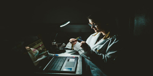 Woman in a lab coat sits at her desk in the dark and is wiping her glasses with a cloth as she does work