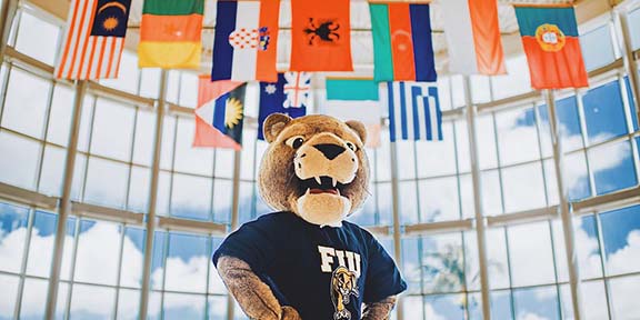 Roary, FIU's mascot, posing for a picture where there's a backdrop of various flags from countries around the globe. 