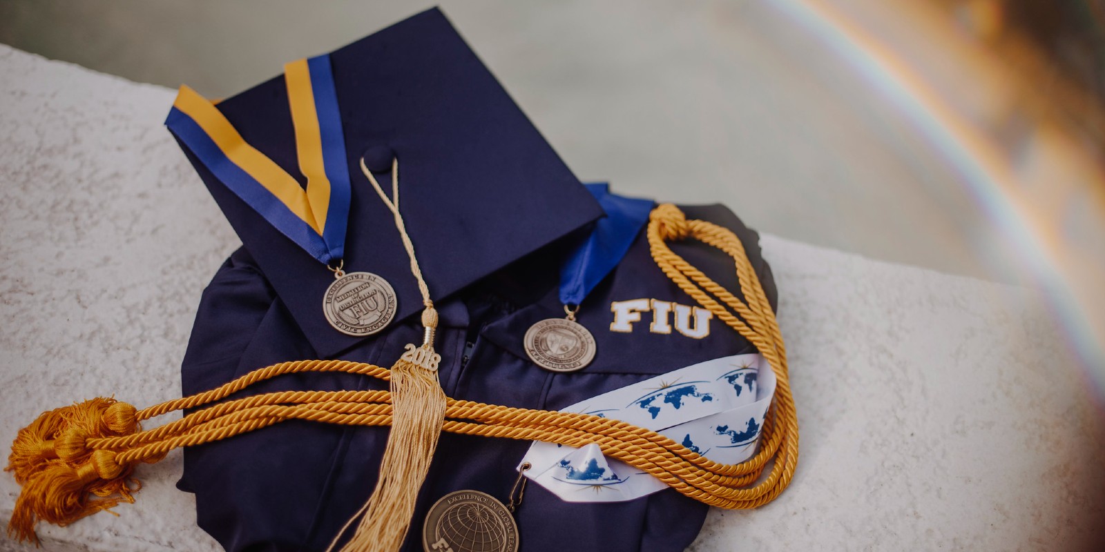 A cap and gown decorated with the FIU gold and blue tassels on a stone bench. 